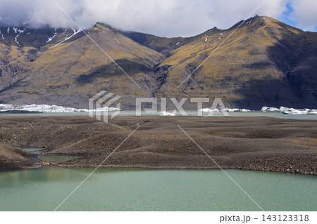 Icebergs beyond gravel shore and turquoise glacial pond beneath mountains in Iceland Icebergs beyond gravel shore and turquoise glacial pond beneath mountains in Iceland 143123318
