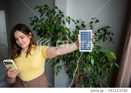 Woman holding a portable solar charger while using smartphone beside indoor greenery. Solar energy green technology and off grid phone charging 143123592