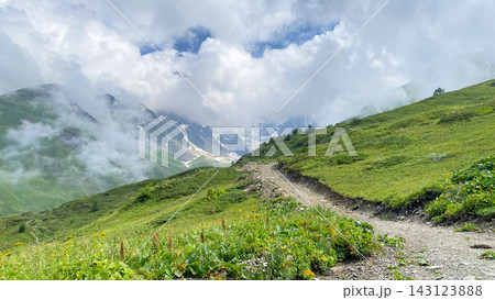 Mountain trail climbs through Svaneti, wide view across green alpine slopes toward cloud-covered Caucasus peaks 143123888