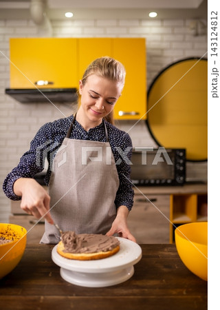 Smiling woman in apron frosting round chocolate cake on turntable in warm modern kitchen 143124812