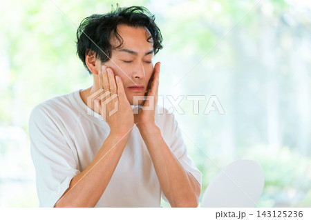 鏡を見ながら頬に手を当てる40代男性。メンズビューティーとエイジングケアのイメージ 鏡を見ながら頬に手を当てる40代男性。メンズビューティーとエイジングケアのイメージ 143125236