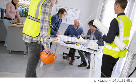 A group of workers and architects stands around a table with drawings and building plans. Discussion of the project budget. A meeting or briefing 143131495