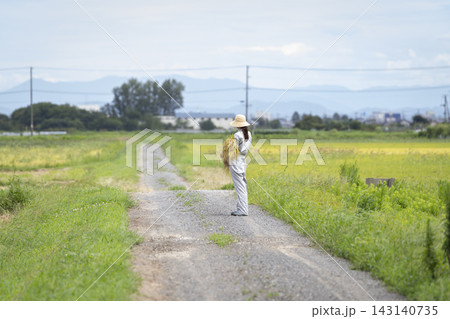 田んぼのあぜ道に立つ稲穂を持った女性の後ろ姿 田んぼのあぜ道に立つ稲穂を持った女性の後ろ姿 143140735