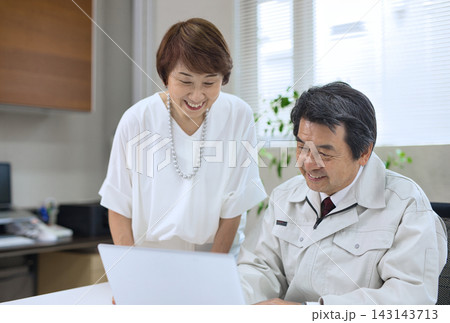 オフィスでパソコンを見ながら会話する作業服の60代男性と60代女性 オフィスでパソコンを見ながら会話する作業服の60代男性と60代女性 143143713