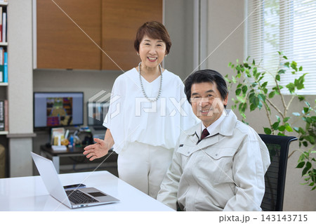オフィスでパソコンを見ながら会話する作業服の60代男性と60代女性 143143715