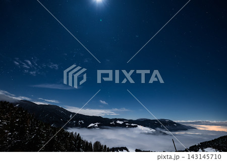 Snow dusted Dolomites peaks rise above a low cloud inversion at night. Clear star filled sky and bright moon cast cool light. Faint village lights suggest remote alpine hotels. 143145710