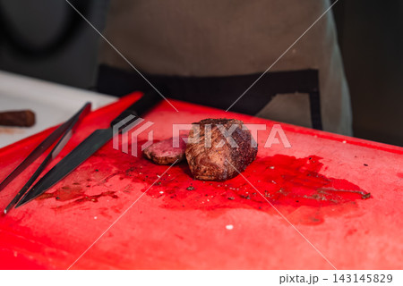 A seared steak rests on a red cutting board with knife and tweezers, juices pooling. Bright task lighting and a chef apron suggest fine dining in Oberlech, Austria. 143145829