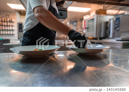 A chef in white sleeve, black apron, and glove plates two dishes on a stainless steel counter in a professional kitchen in Oberlech, Austria, during service. A chef in white sleeve, black apron, and glove plates two dishes on a stainless steel counter in a professional kitchen in Oberlech, Austria, during service. 143145880