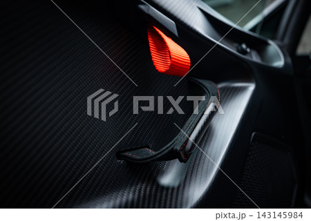 Oblique view shows carbon fiber door panel, pull strap, and red release loop. Soft moody light reveals weave and motorsport cues in the Italian cockpit. Oblique view shows carbon fiber door panel, pull strap, and red release loop. Soft moody light reveals weave and motorsport cues in the Italian cockpit. 143145984