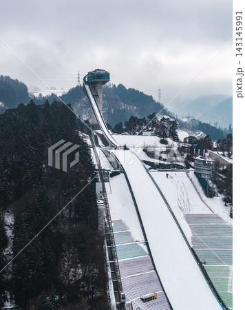 Aerial view of Bergisel Ski Jump in Innsbruck, Austria, with Zaha Hadid tower, long white inrun, stepped landing, winter snow, evergreen forests, and misty Alpine ridges. Aerial view of Bergisel Ski Jump in Innsbruck, Austria, with Zaha Hadid tower, long white inrun, stepped landing, winter snow, evergreen forests, and misty Alpine ridges. 143145991