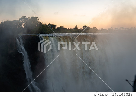 Wide view of Victoria Falls at sunrise shows torrents over a basalt cliff, mist rising like smoke that thunders, treeline backlit, soft light and slight haze present. 143146025