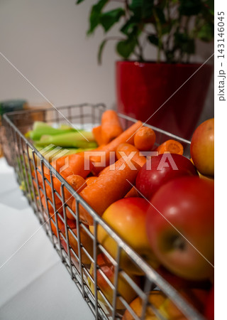 A metal wire basket holds carrots, red and yellow apples, and celery on a white countertop in Oberlech, Austria. A red planter and houseplant sit in soft indoor light. A metal wire basket holds carrots, red and yellow apples, and celery on a white countertop in Oberlech, Austria. A red planter and houseplant sit in soft indoor light. 143146045