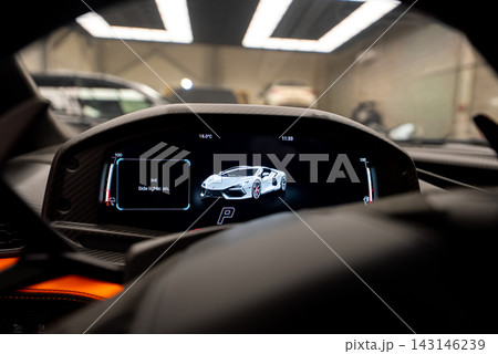 Driver seat view of Lamborghini Revuelto digital cluster shows side lights on, gear P, time 11.33, 18.0 degrees, carbon fiber trim, orange accents, workshop setting. 143146239