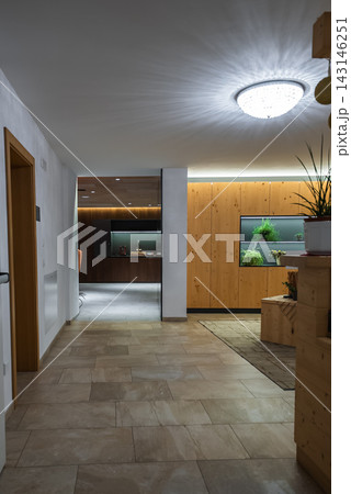 A warm hotel corridor in the Dolomites, Italy leads to a lounge. Light stone tile and paneled wood with plant niches appear under soft ceiling and LED lighting. 143146251