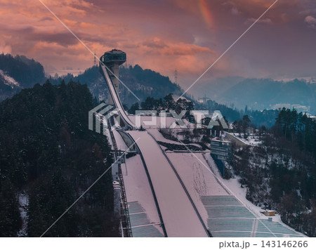 Aerial view of Bergisel Ski Jump in Innsbruck, Austria at dusk, Zaha Hadid tower above snow forests and terraced slopes, faint rainbow over misty Tyrolean mountains. Aerial view of Bergisel Ski Jump in Innsbruck, Austria at dusk, Zaha Hadid tower above snow forests and terraced slopes, faint rainbow over misty Tyrolean mountains. 143146266