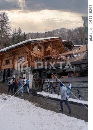Zermatt, Switzerland street shows skiers and pedestrians by a rustic chalet with stone base, carved balconies, ski racks, and terrace lights under misty forested hills. Zermatt, Switzerland street shows skiers and pedestrians by a rustic chalet with stone base, carved balconies, ski racks, and terrace lights under misty forested hills. 143146268