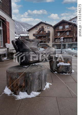 A carved wooden hiking boot on log pedestals stands in a winter plaza in Zermatt, Switzerland, with alpine chalets and snowy mountains under soft overcast daylight. 143146275