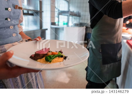 A server in Alpine attire presents seared meat with broccoli, carrots, and puree in a luxury hotel kitchen in Oberlech, Austria, under warm directional light. 143146279
