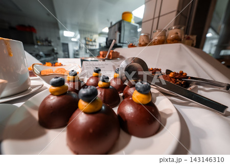 Close up dessert station in Oberlech, Austria shows glossy chocolate domes with saffron cream and blueberries, on a white plate with tongs, warm lights, shallow depth of field. Close up dessert station in Oberlech, Austria shows glossy chocolate domes with saffron cream and blueberries, on a white plate with tongs, warm lights, shallow depth of field. 143146310