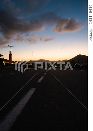 Low angle twilight road scene on Lanzarote, Canary Islands, with silhouetted volcanic hills, streetlights, parked cars, and one approaching headlight under a dramatic sky. 143146456
