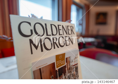 A menu titled Goldener Morgen sits on a table in Hotel Goldener Berg, Oberlech, Austria. Warm red seating, curtained windows, muted lighting, and shallow depth of field are visible. A menu titled Goldener Morgen sits on a table in Hotel Goldener Berg, Oberlech, Austria. Warm red seating, curtained windows, muted lighting, and shallow depth of field are visible. 143146467
