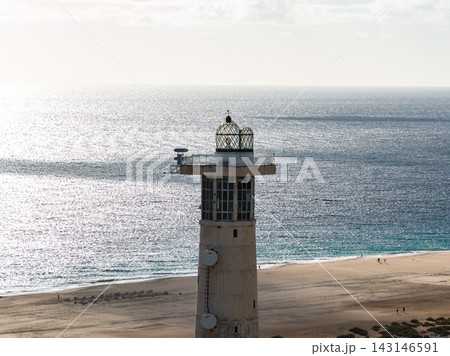 Morro Jable Lighthouse stands by a wide beach in Fuerteventura, Spain. Afternoon light glints on Atlantic waters as a few people stroll near the waterline. Morro Jable Lighthouse stands by a wide beach in Fuerteventura, Spain. Afternoon light glints on Atlantic waters as a few people stroll near the waterline. 143146591