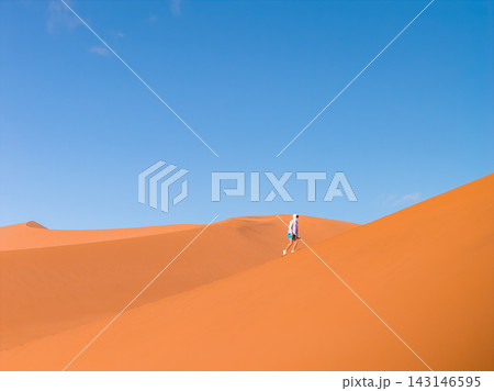 A lone hiker climbs a steep dune in Sossusvlei's Deadvlei, Namibia. Early or late light defines crisp contours and long soft shadows under a cloudless sky. 143146595
