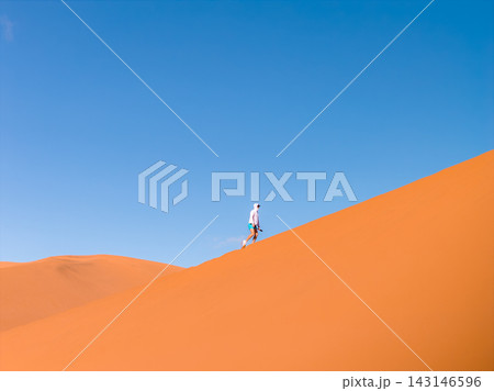 A lone hiker ascends a steep sand dune in Sossusvlei, Namibia. The person wears sun gear and carries a camera. Strong contrast, clean lines, and wide negative space show scale. 143146596