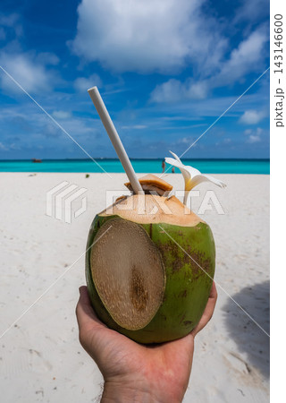 A hand holds Maldives coconut with blank name space with frangipani and straw, set before white sand and a turquoise lagoon under a bright sky, captured in daylight focus. 143146600