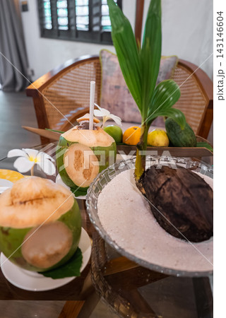 Two Coco Palm coconuts with straws sit by a sprouting coconut seedling in a shallow sand bowl, inside a Maldives beach villa lounge with rattan and shuttered windows. Two Coco Palm coconuts with straws sit by a sprouting coconut seedling in a shallow sand bowl, inside a Maldives beach villa lounge with rattan and shuttered windows. 143146604