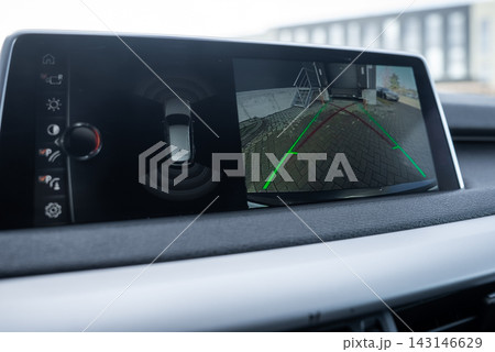 Dashboard screen shows BMW iDrive menu, top down sensor graphic, and rear view with wall, metal bike rack, and potted plants on a cobblestone driveway in daytime. Dashboard screen shows BMW iDrive menu, top down sensor graphic, and rear view with wall, metal bike rack, and potted plants on a cobblestone driveway in daytime. 143146629