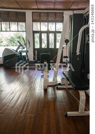 Polished wooden flooring, cable machine with bench, leg extension, and elliptical sit in soft daylight. Louvered windows and glass doors reveal tropical plants in a Maldives resort. Polished wooden flooring, cable machine with bench, leg extension, and elliptical sit in soft daylight. Louvered windows and glass doors reveal tropical plants in a Maldives resort. 143146669