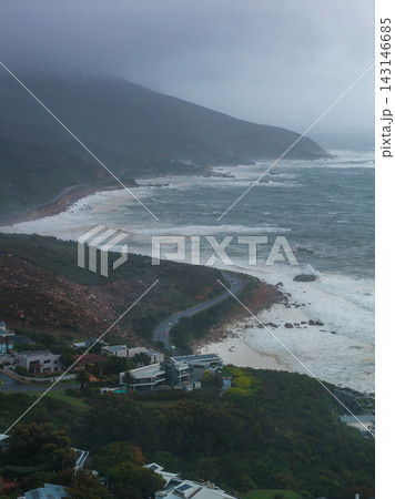 Aerial coastal scene in Cape Town shows a winding cliff road, stormy Atlantic surf, low clouds on a dark ridge, modern houses, green vegetation, and scattered boulders. Aerial coastal scene in Cape Town shows a winding cliff road, stormy Atlantic surf, low clouds on a dark ridge, modern houses, green vegetation, and scattered boulders. 143146685