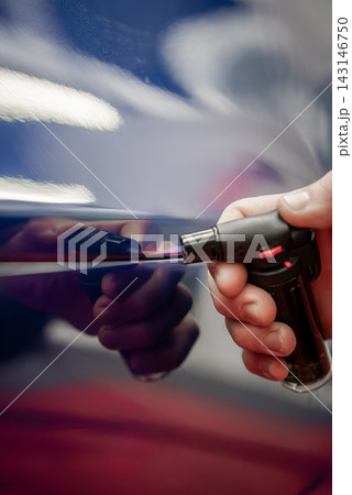 A technician uses a butane micro torch on a thin line of a glossy blue car door, with reflections, bright shop lighting, bokeh highlights, and shallow depth of field. 143146750