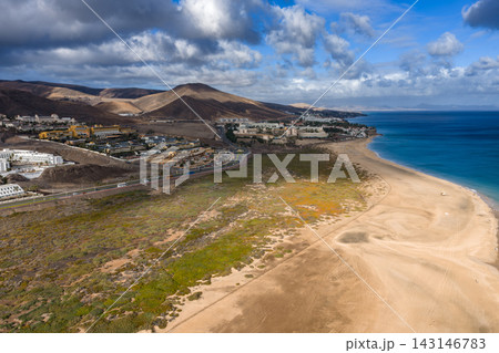 Aerial scene shows Morro Jable on Fuerteventura, Canary Islands. Turquoise water meets long sands, resort complexes, volcanic hills, tidal plain, and moving cloud shadows. 143146783