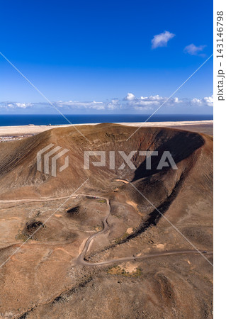 Aerial view of a rust toned caldera, badlands, and Atlantic shore in Fuerteventura. A winding dirt track crosses arid terrain under clear midday light and deep skies. 143146798