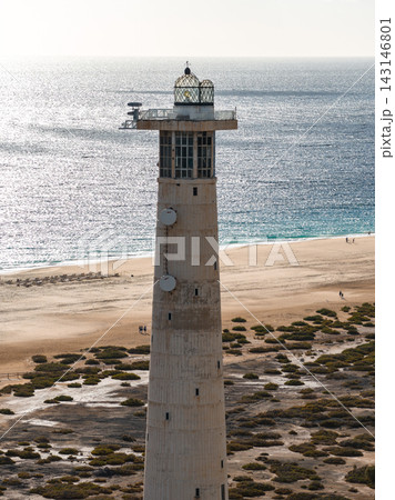 Aerial view shows Morro Jable Lighthouse in Fuerteventura, Canary Islands. Cylindrical shaft and glass lantern stand by salt flats, beachgoers, and silvery Atlantic. 143146801