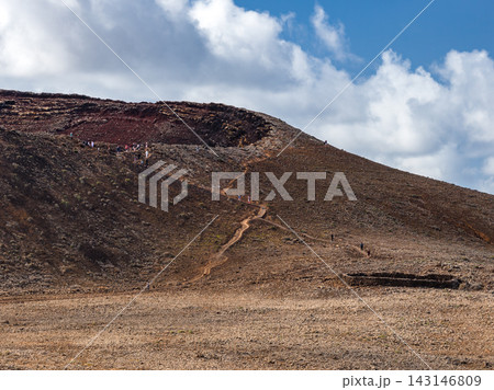 An arid volcanic hillside rises under a bright sky in Fuerteventura. A winding trail leads hikers toward a crater rim with dark red lava rock in crisp midday light. An arid volcanic hillside rises under a bright sky in Fuerteventura. A winding trail leads hikers toward a crater rim with dark red lava rock in crisp midday light. 143146809