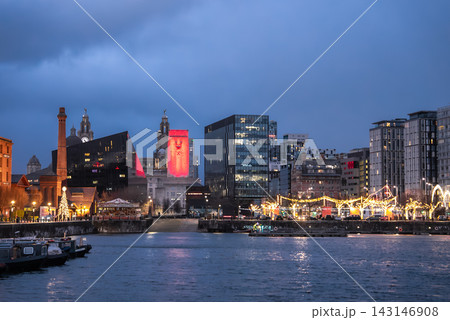 Royal Albert Dock in Liverpool at twilight, clock towers of the Royal Liver Building and a tall red lit tower stand beyond, Christmas lights and market stalls line the quay. Royal Albert Dock in Liverpool at twilight, clock towers of the Royal Liver Building and a tall red lit tower stand beyond, Christmas lights and market stalls line the quay. 143146908