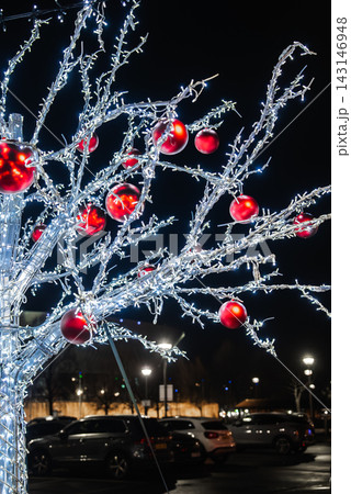 A festive artificial tree with white LED lights and red baubles stands in a Liverpool car park at night, UK plates visible, cool lights against a black sky. A festive artificial tree with white LED lights and red baubles stands in a Liverpool car park at night, UK plates visible, cool lights against a black sky. 143146948
