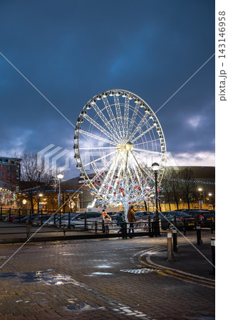 Wheel of Liverpool near M and S Bank Arena rises over wet cobblestones as people gather by a Christmas tree with oversized red baubles during a festive winter evening. Wheel of Liverpool near M and S Bank Arena rises over wet cobblestones as people gather by a Christmas tree with oversized red baubles during a festive winter evening. 143146958