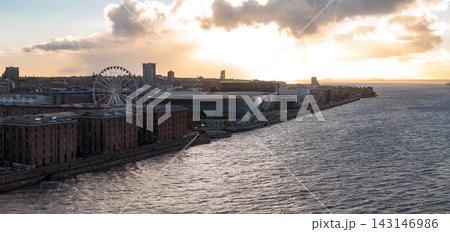 Aerial view at sunrise shows Liverpool, UK, the River Mersey, the Ferris wheel, ACC Liverpool and M and S Bank Arena, and Albert Dock warehouses under warm light and clouds. Aerial view at sunrise shows Liverpool, UK, the River Mersey, the Ferris wheel, ACC Liverpool and M and S Bank Arena, and Albert Dock warehouses under warm light and clouds. 143146986