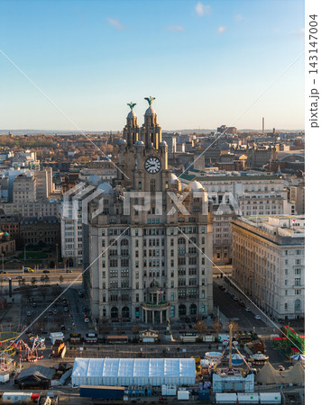 Aerial sunrise view of Liverpool, UK, with the Royal Liver Building, clock face, and Liver Bird statues. Pier Head surrounds, fairground tents animate streets, River Mersey gleams. 143147004