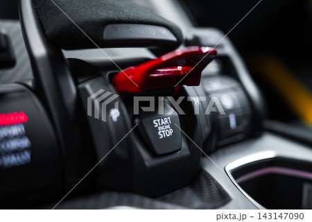 A tight close up shows the red flip cover over the START STOP button on a Lamborghini Urus Performante center console, with P and M S selectors and carbon fiber trim. A tight close up shows the red flip cover over the START STOP button on a Lamborghini Urus Performante center console, with P and M S selectors and carbon fiber trim. 143147090