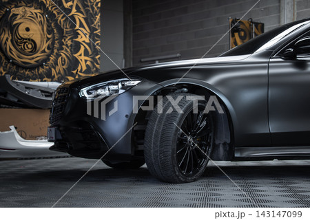 A matte black Mercedes Benz S Class sits indoors with wheel turned outward, showing black multi spoke rims and performance brakes, tools and a gold motif mural in view. 143147099