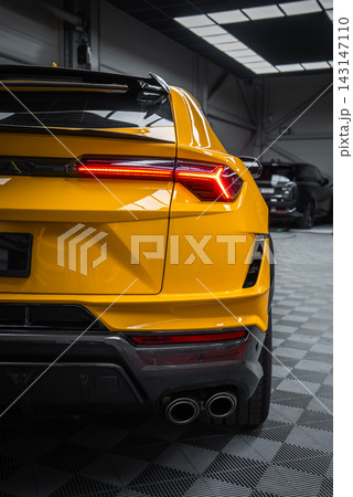 Rear three quarter view shows angular LED taillight, dual exhausts, and carbon fiber diffuser under soft overhead light, with a dark sedan in a showroom setting. 143147110