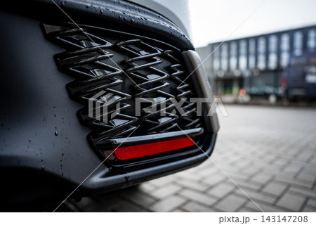A close up shows the Lexus RX 450h plus rear bumper, glossy black lattice vent, and slim red reflector. Raindrops sit on dark paint near blurred buildings and parked cars. 143147208