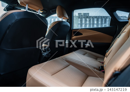 Close up interior shows tan perforated leather seats, black panels, and rear vents. Shot from left rear door with raindrops on windows and a blurred modern building. Close up interior shows tan perforated leather seats, black panels, and rear vents. Shot from left rear door with raindrops on windows and a blurred modern building. 143147219