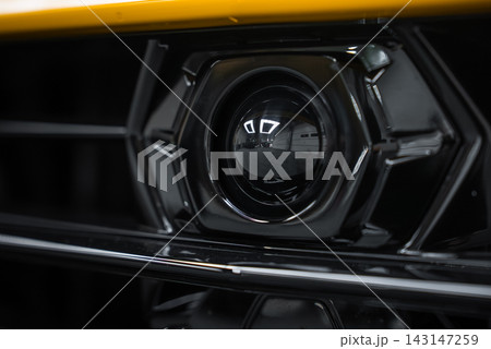 An extreme close up shows the front camera in a yellow Lamborghini Urus Performante grille, its glossy hexagonal housing reflecting ceiling lights in an indoor studio. An extreme close up shows the front camera in a yellow Lamborghini Urus Performante grille, its glossy hexagonal housing reflecting ceiling lights in an indoor studio. 143147259