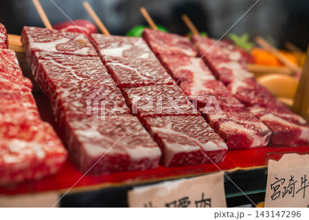 Marbled wagyu skewers line a red tray at a Kyoto market stall, with handwritten Japanese labels. Warm natural light and shallow focus emphasize texture and craft. 143147296